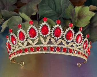 May include: A gold-toned tiara featuring a series of vibrant red teardrop-shaped crystals arranged along the top edge, accented by smaller round red crystals along the base and clear rhinestones throughout the intricate metal leaf-like frame.
