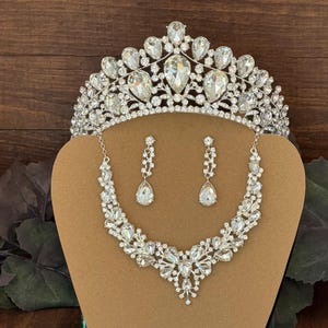 May include: A silver-toned jewelry set consisting of a tiara, a necklace, and a pair of drop earrings. Each piece is encrusted with clear, faceted crystals in various shapes, including pear and round cuts, set in a metallic frame.
