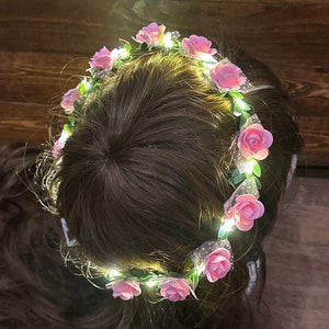May include: A pink flower crown with lights, worn on a person's head. The crown is made of artificial flowers and has a wire frame. The lights are white and are strung around the flowers.