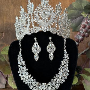 May include: A silver tiara with clear rhinestones, a matching necklace, and a pair of earrings. The jewelry set is displayed on a black stand.