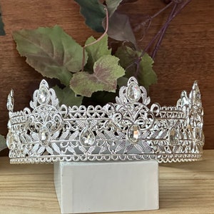 May include: Silver crown with intricate detailing and clear rhinestones. The crown is on a white pedestal.