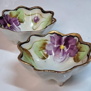 Peut inclure: Deux petits bols blancs à bords festonnés avec des fleurs violettes et des feuilles vertes peintes à la main. Les bords sont dorés. Chaque bol a trois petits pieds.
