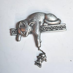 Peut inclure: Broche argentée en forme de chat au design ludique. Le chat est allongé sur une barre décorative, une patte tendue vers une petite souris. La broche est en métal et présente une surface texturée et détaillée. Un accessoire charmant pour les amoureux des chats.
