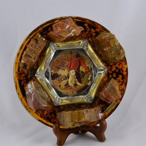 May include: A round, amber-colored ashtray with a picture of a person and a dog in the center. The ashtray is decorated with several pieces of red, orange, and green stone. The text "Petrified Wood" is visible on the bottom right side of the ashtray.