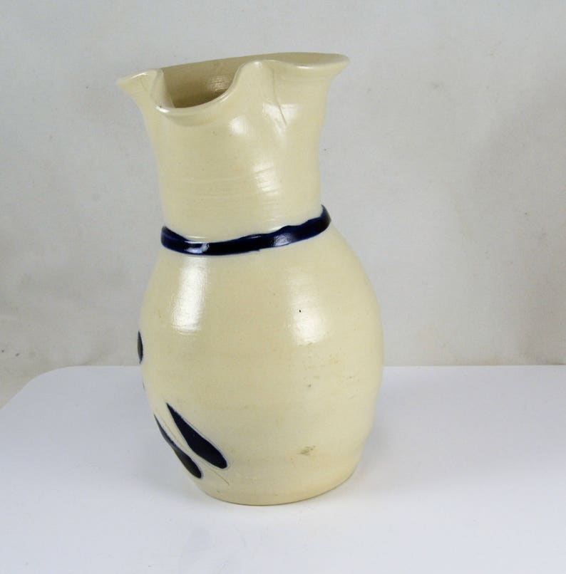 May include: A white ceramic pitcher with a blue band around the neck and blue painted designs. The pitcher has a wide mouth and a slightly curved body.