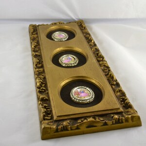 May include: A gold-framed wall decor with three round, framed, porcelain medallions featuring romantic scenes. The medallions are set against a dark brown background.