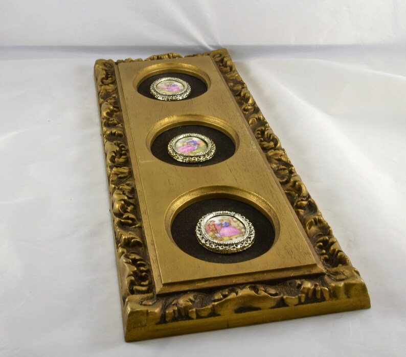 May include: A gold-framed wall decor with three round, framed, porcelain medallions featuring romantic scenes. The medallions are set against a dark brown background.