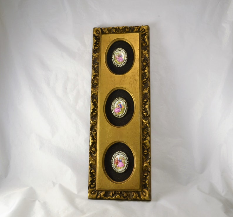 May include: A gold framed wall decor with three round porcelain medallions featuring images of people in vintage clothing.