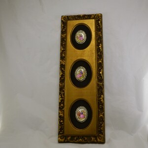May include: A gold framed wall decor with three oval-shaped black and gold frames. Each frame contains a small porcelain cameo with a couple in a romantic setting.