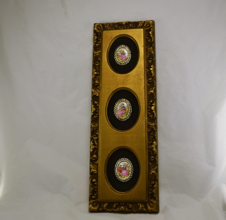 May include: A gold framed wall decor with three oval-shaped black and gold frames. Each frame contains a small porcelain cameo with a couple in a romantic setting.