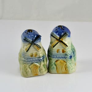 May include: A pair of ceramic salt and pepper shakers shaped like windmills. The shakers are painted in shades of blue, green, and yellow, with white accents. The windmills have white blades and brown roofs.
