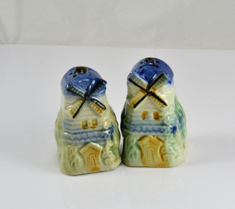 May include: A pair of ceramic salt and pepper shakers shaped like windmills. The shakers are painted in shades of blue, green, and yellow, with white accents. The windmills have white blades and brown roofs.