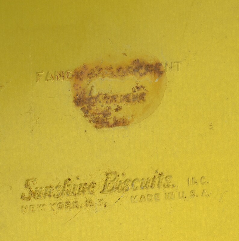 K&ouml;nnte beinhalten: Eine gelbe Metalldose mit dem Text "Sunshine Biscuits, Inc. New York, N.Y. Made in U.S.A." auf der Unterseite. Die Oberseite der Dose hat einen verblassten, rostigen Aufkleber mit dem Text "Fancy Crackers".