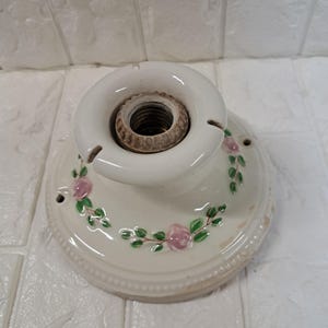 Peut inclure: Une base de luminaire ronde en c&eacute;ramique blanc cass&eacute; avec une douille m&eacute;tallique filet&eacute;e centrale. La surface est orn&eacute;e d'un motif peint de petites roses roses et de feuilles vertes dispos&eacute;es en une guirlande circulaire autour du centre.