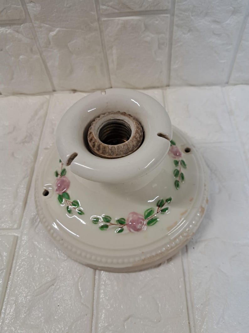 Peut inclure: Une base de luminaire ronde en c&eacute;ramique blanc cass&eacute; avec une douille m&eacute;tallique filet&eacute;e centrale. La surface est orn&eacute;e d'un motif peint de petites roses roses et de feuilles vertes dispos&eacute;es en une guirlande circulaire autour du centre.