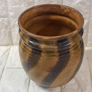 May include: A handcrafted, round ceramic pot with a wide opening. The pot features a brown and black striped pattern around the exterior. The interior is a darker brown. The pot is likely used for decorative purposes or for holding plants.