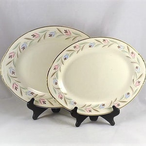 May include: Two cream-colored oval and round serving platters with a floral border in pink, blue, and green, and gold trim. The platters are displayed on black stands. The vintage dishware is likely for serving food.