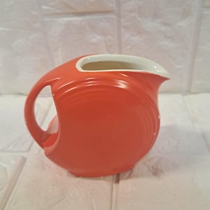May include: A vibrant orange ceramic pitcher with a curved handle and a white interior rim. The pitcher has a smooth, glossy finish and a classic design, perfect for serving beverages. The pitcher measures approximately 7 inches in height.