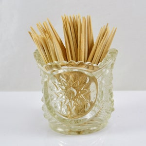 May include: A clear glass toothpick holder with a starburst design. The holder is filled with wooden toothpicks.