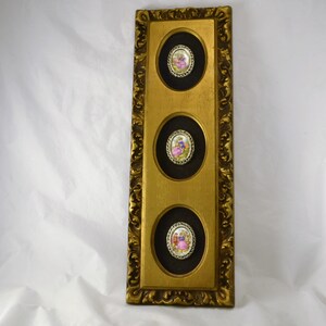 May include: A gold framed wall art piece with three oval-shaped portraits of people in vintage clothing. The portraits are set against a black background and are surrounded by a gold trim.
