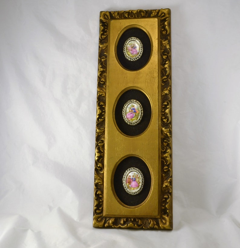 May include: A gold framed wall art piece with three oval-shaped portraits of people in vintage clothing. The portraits are set against a black background and are surrounded by a gold trim.
