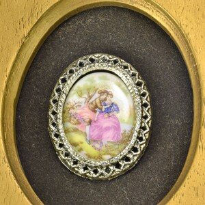 May include: A framed oval porcelain cameo with a delicate silver filigree border. The cameo depicts a couple in a romantic embrace in a garden setting.