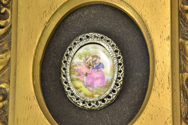 May include: A framed oval porcelain cameo with a delicate silver filigree border. The cameo depicts a couple in a romantic embrace in a garden setting.