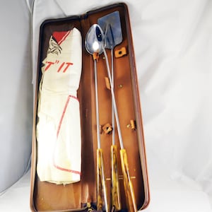 May include: A brown leather case with a zipper closure, containing a set of four grilling tools with wooden handles. The tools include a spatula, a spoon, and two long skewers. The case also includes a white cloth with the text "T'IT" printed in red.