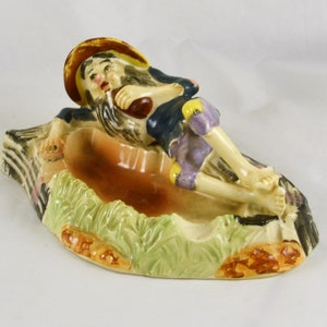 May include: A ceramic ashtray featuring a whimsical figure of a man in a straw hat and shorts, reclining on a log with a bottle in hand. The ashtray is decorated with green grass and brown rocks.