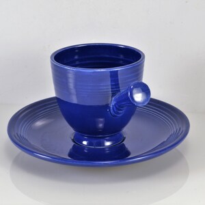 May include: A blue ceramic teacup and saucer set. The teacup has a unique handle that resembles a knob. The saucer has a scalloped edge.