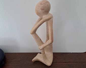 May include: A hand-carved wooden sculpture of a person in a seated pose. The sculpture is made of light brown wood and has a smooth, polished finish.