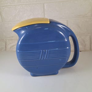 May include: A blue ceramic pitcher with a yellow lid. The pitcher has a rounded shape and a handle on the side. The pitcher has a design of horizontal lines and the letter "N" on the front.