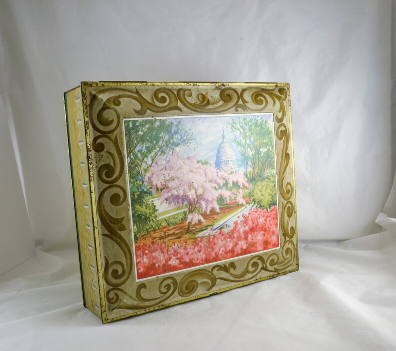 K&ouml;nnte beinhalten: Vintage Blechdose mit einem verzierten goldenen und wei&szlig;en Rahmen und einem farbenfrohen Bild des US-Kapitols mit Kirschbl&uuml;ten im Vordergrund.