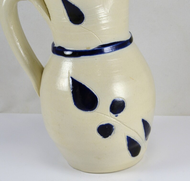 May include: A white ceramic pitcher with a blue rim and blue leaf designs. The pitcher has a handle and a rounded body.