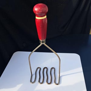 May include: A red and gold potato masher with a wooden handle. The masher has a wavy, metal base.