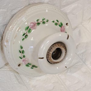 Peut inclure: Un socle de plafonnier en c&eacute;ramique cr&egrave;me de forme ronde, orn&eacute; d'un motif floral peint &agrave; la main repr&eacute;sentant des roses roses et des feuilles vertes. Le luminaire poss&egrave;de une douille m&eacute;tallique centrale filet&eacute;e et trois trous de fixation.
