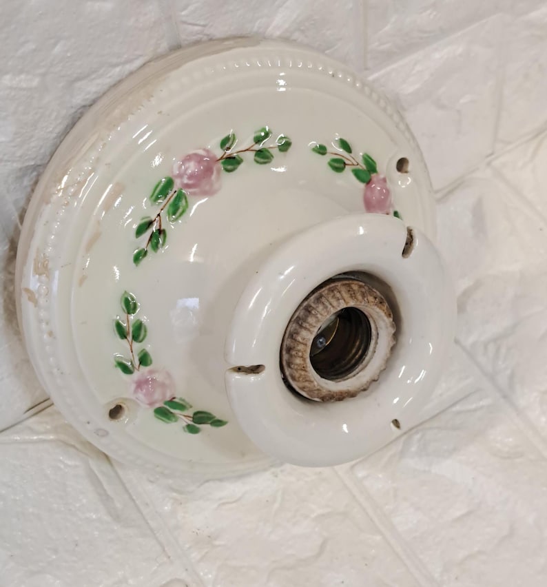 Peut inclure: Un socle de plafonnier en c&eacute;ramique cr&egrave;me de forme ronde, orn&eacute; d'un motif floral peint &agrave; la main repr&eacute;sentant des roses roses et des feuilles vertes. Le luminaire poss&egrave;de une douille m&eacute;tallique centrale filet&eacute;e et trois trous de fixation.