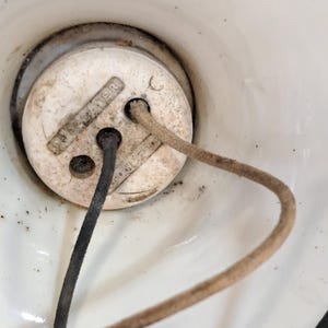 Peut inclure: Une base de douille &eacute;lectrique en c&eacute;ramique blanche de forme circulaire, comportant deux trous o&ugrave; sont ins&eacute;r&eacute;s un fil noir et un fil recouvert de tissu beige. La surface du composant en c&eacute;ramique blanche pr&eacute;sente des traces de poussi&egrave;re.