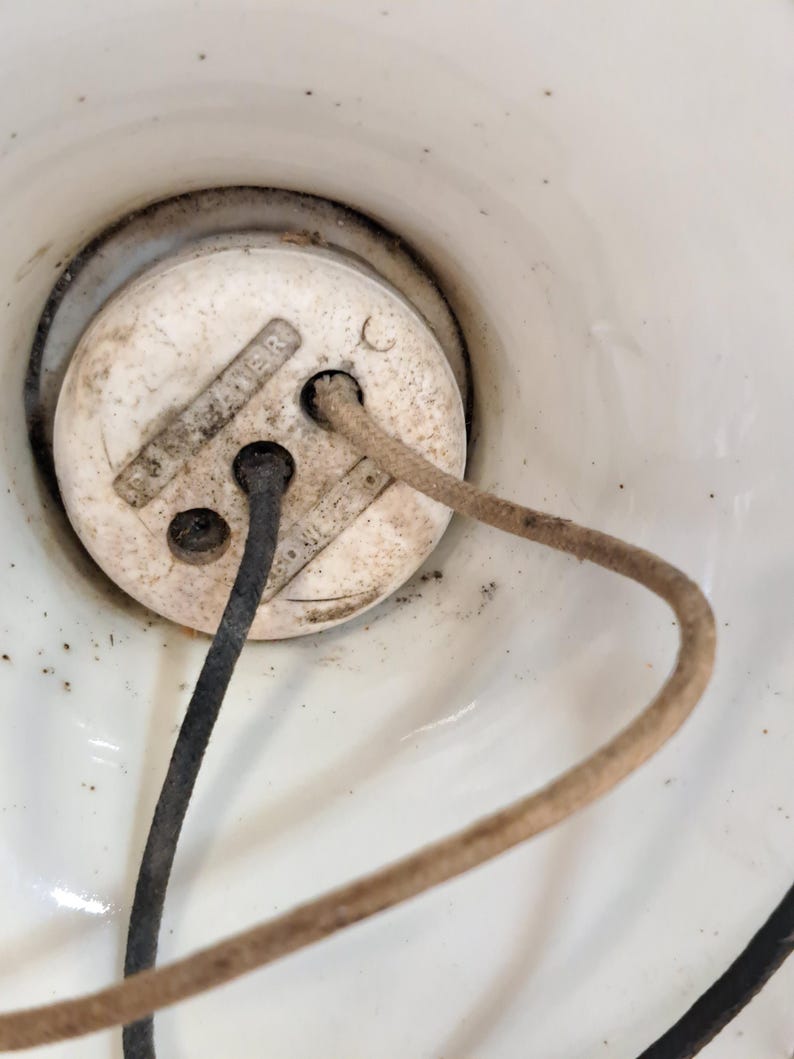 Peut inclure: Une base de douille &eacute;lectrique en c&eacute;ramique blanche de forme circulaire, comportant deux trous o&ugrave; sont ins&eacute;r&eacute;s un fil noir et un fil recouvert de tissu beige. La surface du composant en c&eacute;ramique blanche pr&eacute;sente des traces de poussi&egrave;re.