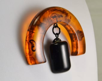 Könnte beinhalten: Eine Vintage-Brosche mit einem großen, halbrunden Harzstück in Bernstein-Farbe und einem schwarzen, rechteckigen Harz-Anhänger, der an einer Metallkette hängt.
