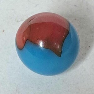May include: A colorful glass marble with a two-tone design. The top half is a vibrant red, transitioning to a deep brown, while the bottom half is a bright, glossy blue. The marble is a classic toy, perfect for collectors or casual play.