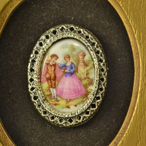 May include: A framed oval cameo with a gold-colored metal frame. The cameo features a painted scene of a couple in a garden setting.