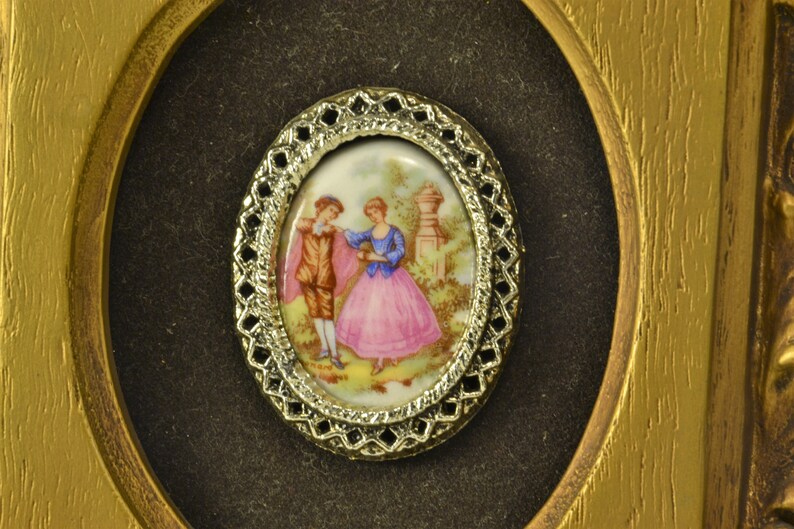 May include: A framed oval cameo with a gold-colored metal frame. The cameo features a painted scene of a couple in a garden setting.
