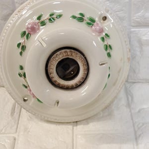 Peut inclure: Une base de plafonnier ronde en c&eacute;ramique blanche orn&eacute;e d'un motif floral peint &agrave; la main repr&eacute;sentant de petites roses roses et des feuilles vertes. Le centre comprend une douille circulaire pour ampoule et le bord est perl&eacute;.
