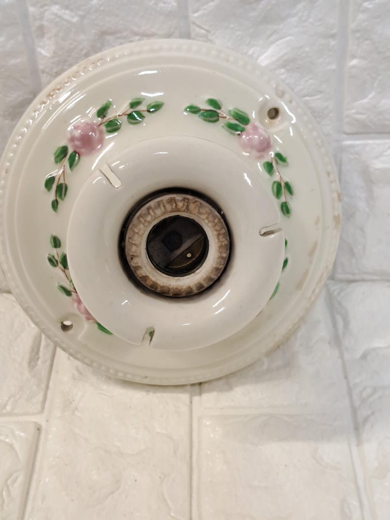 Peut inclure: Une base de plafonnier ronde en c&eacute;ramique blanche orn&eacute;e d'un motif floral peint &agrave; la main repr&eacute;sentant de petites roses roses et des feuilles vertes. Le centre comprend une douille circulaire pour ampoule et le bord est perl&eacute;.