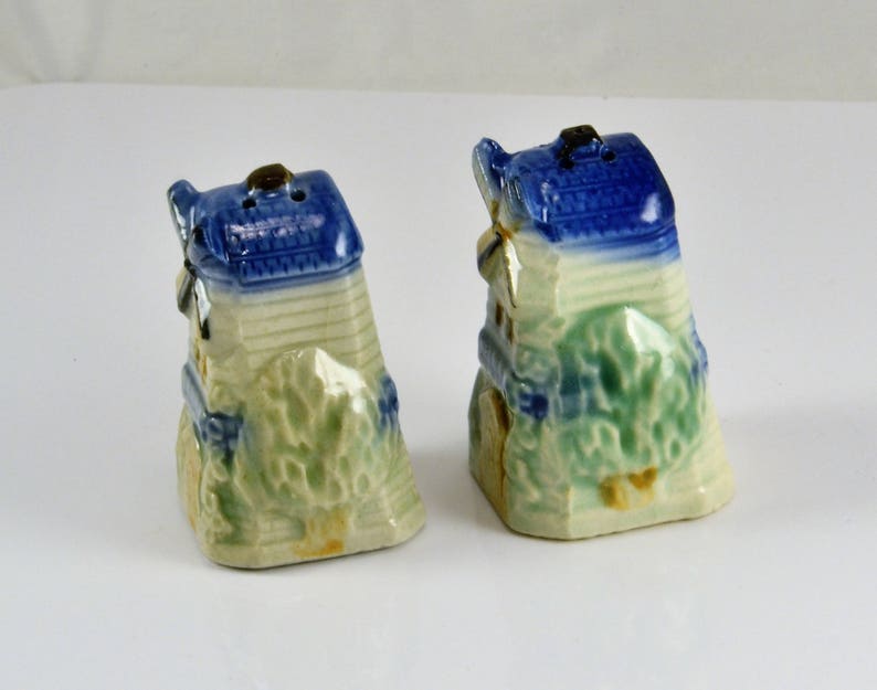 May include: A pair of ceramic salt and pepper shakers shaped like houses with blue roofs and green trees. The shakers are hand-painted and have a rustic charm.