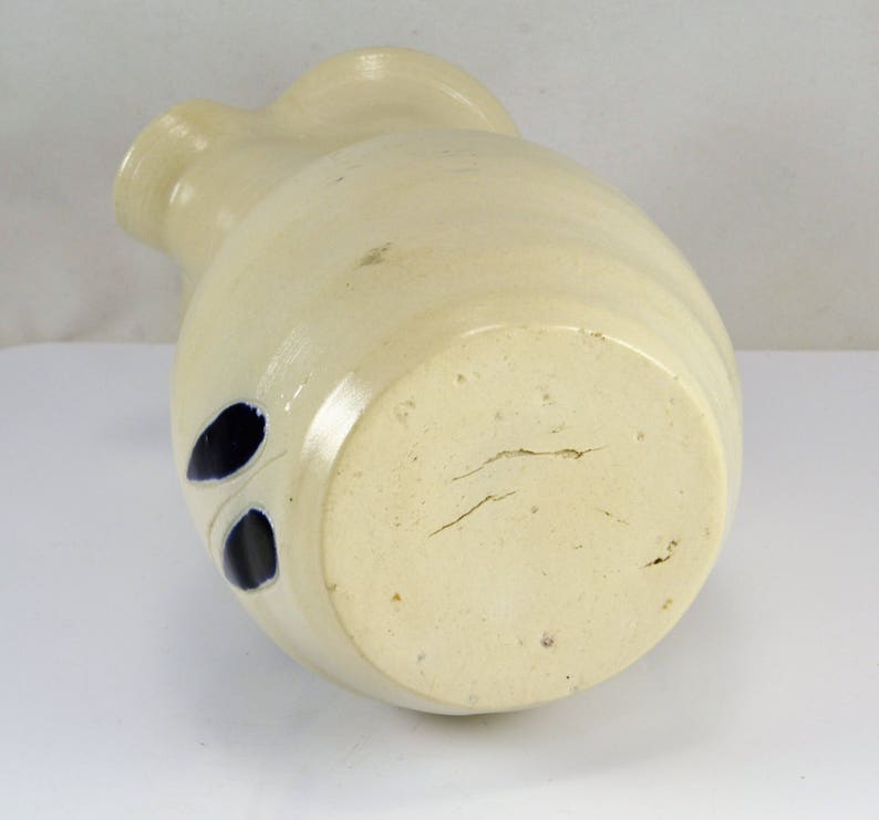 May include: A white ceramic pitcher with blue leaf designs. The pitcher has a rounded body and a wide mouth. The bottom of the pitcher is unglazed and shows signs of wear.