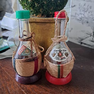 May include: Two small, vintage glass bottles with green and red caps. The bottles are wrapped in woven twine and have labels that read "Italian Swiss Colony". The bottles sit on a brown surface.