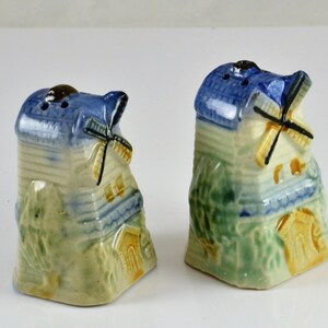 May include: A pair of ceramic salt and pepper shakers shaped like windmills. The shakers are painted in shades of blue, green, and yellow, with white accents. The windmills have working blades.