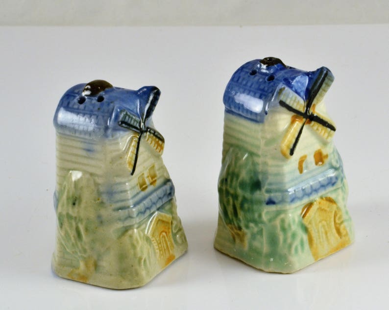 May include: A pair of ceramic salt and pepper shakers shaped like windmills. The shakers are painted in shades of blue, green, and yellow, with white accents. The windmills have working blades.